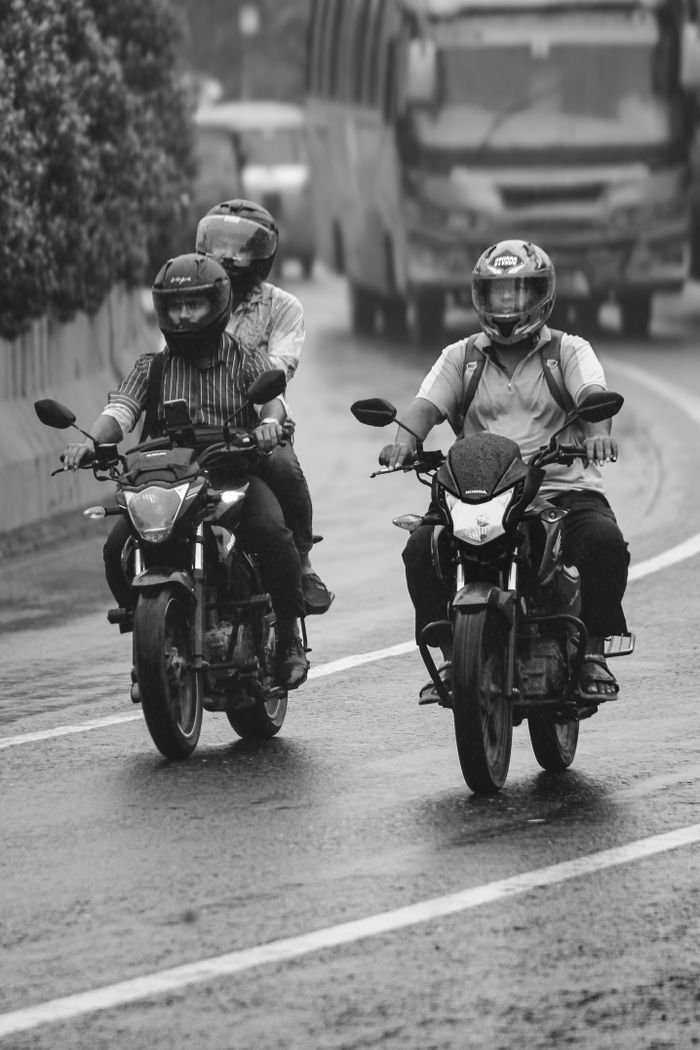 Three motorcyclists wearing helmets ride through a wet urban street in black and white.